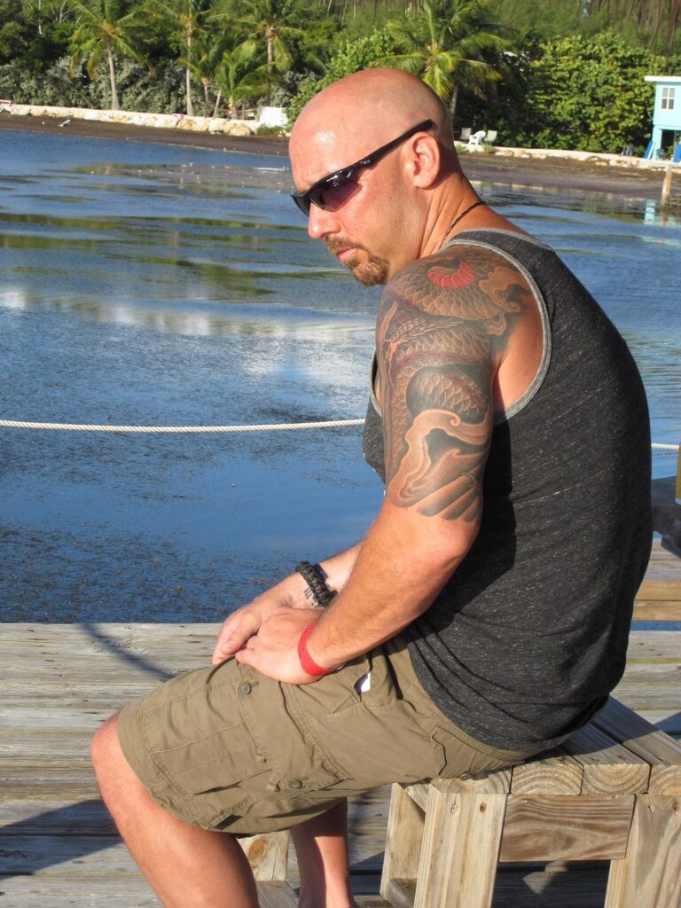 Shayne standing on a wooden lake dock, shirtless, showing a full dragon sleeve tattoo on his right arm.