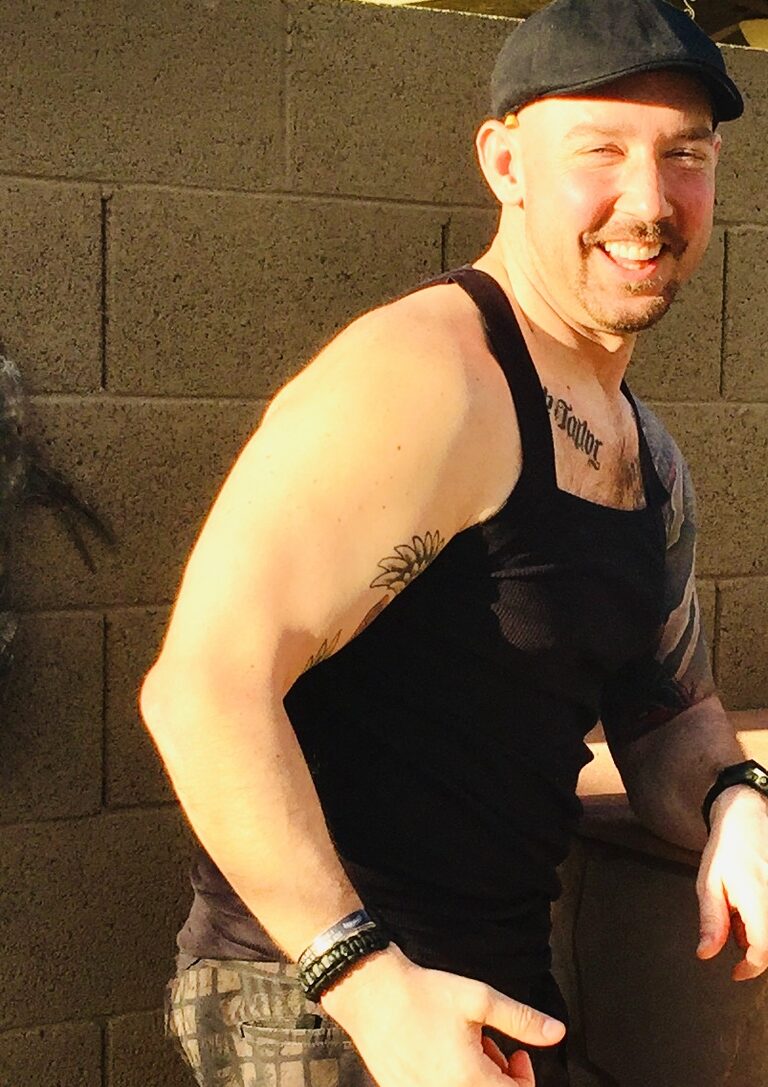 Close-up of Shayne smiling at the camera, his daughter's name 'Taylor' tattooed across his chest.